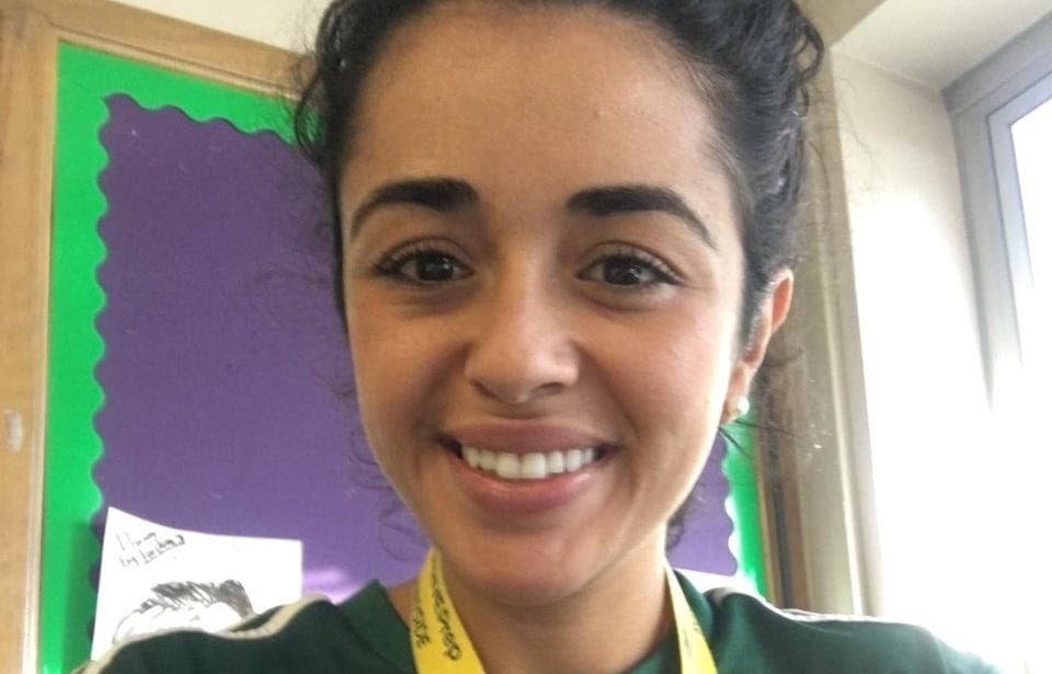 A selfie of Wendy Gutierrez. She is smiling in her classroom, wearing an ID lanyard and a green T-shirt in front of a purple bulletin board 