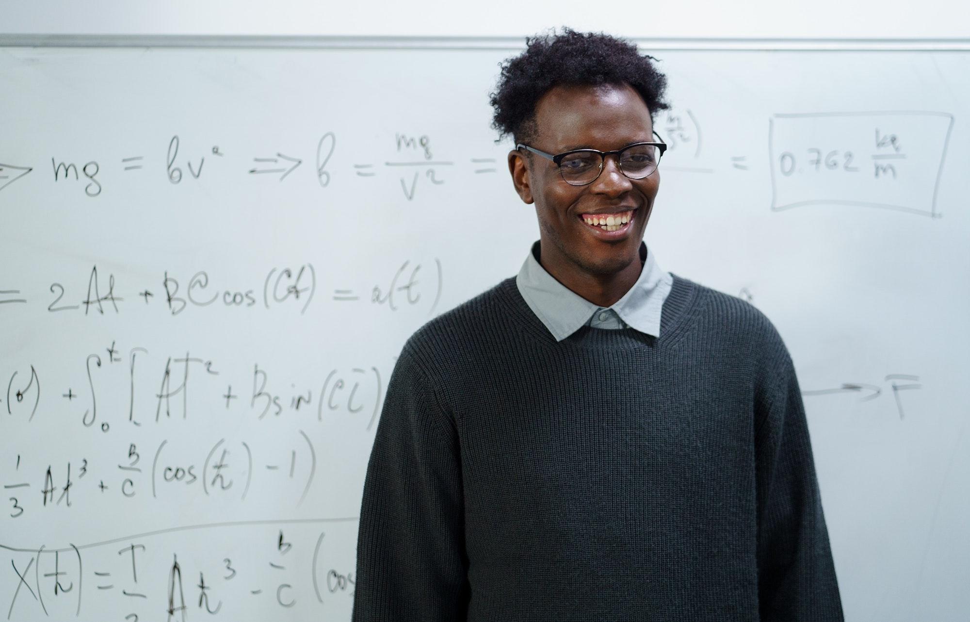 A math teacher in a Colorado public school stands in front of the whiteboard in his classroom. He wears glasses and is looking at the camera and smiling. There are math equations filling up the whiteboard behind him.