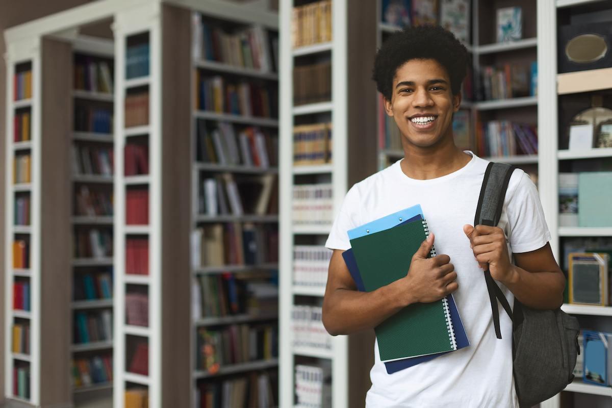 A future Colorado teacher standing in his campus library. He’s wearing a white T-shirt and holding books in one hand and a backpack on his shoulder with the other. 